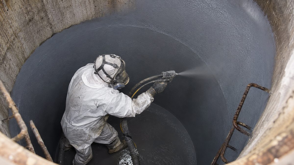 Worker spray-applying polyurea protective lining inside a concrete manhole and sewer